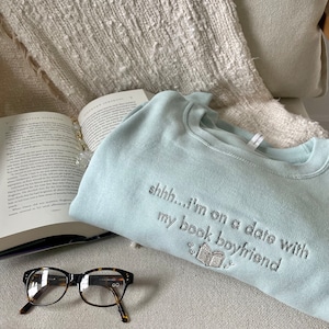 Peut inclure: Un sweat-shirt bleu clair avec le texte "shhh...i'm on a date with my book boyfriend" brod&eacute; sur le devant. Le sweat-shirt est pos&eacute; sur une surface blanche et moelleuse avec une paire de lunettes brunes et un livre.