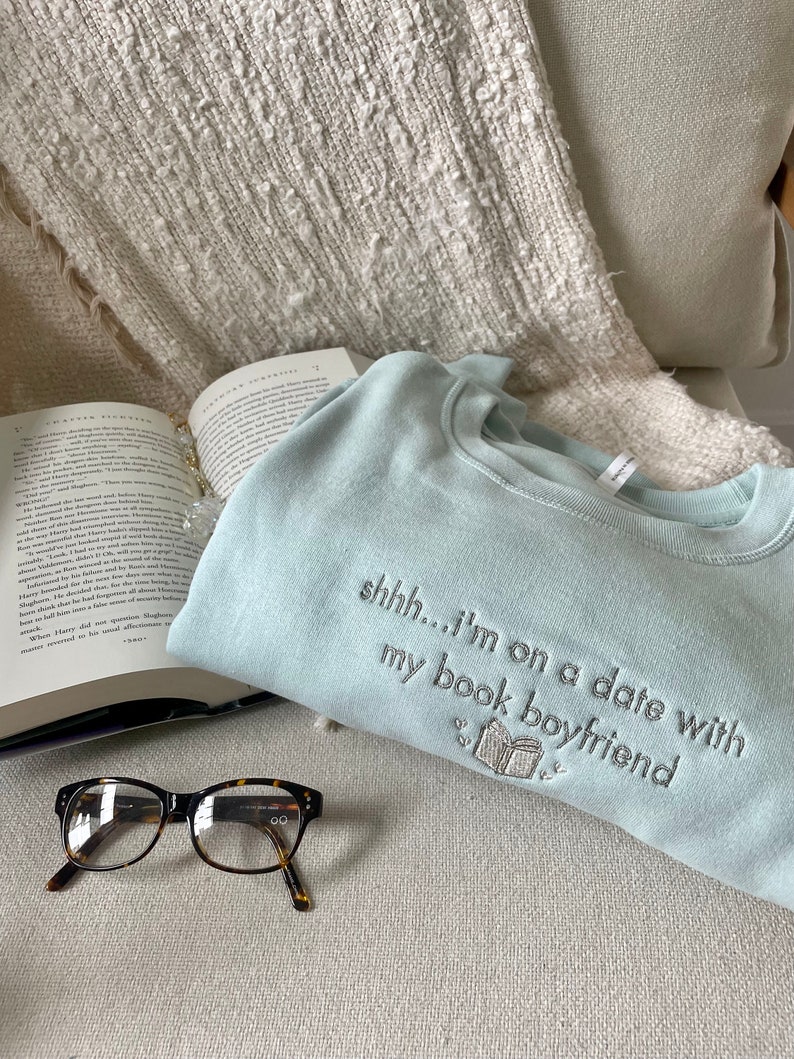 Peut inclure: Un sweat-shirt bleu clair avec le texte "shhh...i'm on a date with my book boyfriend" brod&eacute; sur le devant. Le sweat-shirt est pos&eacute; sur une surface blanche et moelleuse avec une paire de lunettes brunes et un livre.