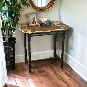 May include: A small, antique wooden side table with a dark brown finish and ornate legs. The tabletop is a lighter brown with floral accents and a gold trim. A framed photo and books rest on the table. The table is approximately 71 cm tall.