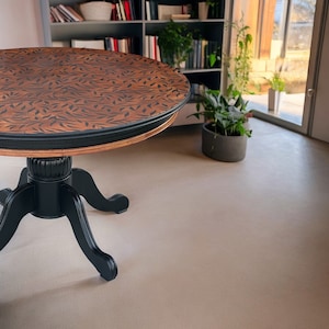 May include: A round wooden table with a dark brown leaf pattern on the tabletop. The table has a black trim and a black pedestal base with three curved legs. The table is approximately 40 inches in diameter.