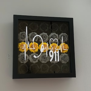 May include: A framed floral arrangement featuring gray and yellow roses. The design includes a white graphic of a heartbeat and the number 911. The frame is black and mounted on a light gray wall.