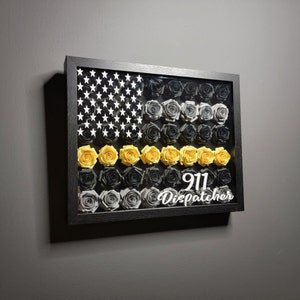 May include: A framed American flag with a black and gray floral design. The flag has a yellow stripe across the middle with the text "911 Dispatcher" below it.