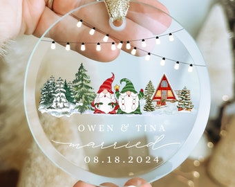 May include: Clear, round Christmas ornament with a gold ribbon. The ornament features a winter scene with two gnomes, a red cabin, and string lights. Text includes "Owen & Tina married 08.18.2024".