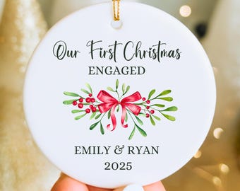 May include: Round white ceramic Christmas ornament with gold hanging string. The ornament reads "Our First Christmas ENGAGED", with the names "EMILY & RYAN 2025". A watercolour design of a red bow and foliage decorates the bottom.