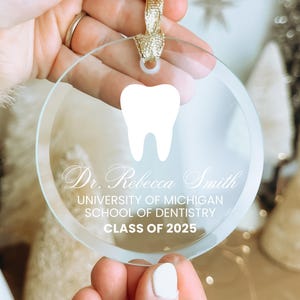 May include: A clear glass ornament with a white tooth design and the text "Dr. Rebecca Smith, University of Michigan School of Dentistry, Class of 2025".