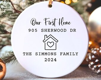 May include: A white, round Christmas ornament with the text "Our First Home," address, a house icon with a heart, "THE SIMMONS FAMILY," and the year 2024. The ornament hangs from a gold string.