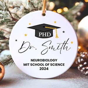May include: White round Christmas ornament with a black graduation cap with the word "PHD" on it. The ornament also has gold stars and the text "Dr. Smith", "Neurobiology", "MIT School of Science", and "2024".
