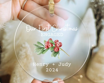 May include: A clear, circular Christmas ornament with a gold ribbon. The ornament has the text "Our First Christmas ENGAGED" and the names "Henry + Judy 2024" with a watercolour sprig of red berries and green leaves.