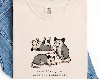 May include: Ivory-colored t-shirt with a graphic of three cartoon opossums, one lying down, one sitting, and one on its back. The text "most likely to meet the deductible" is below the opossums. The shirt is folded on top of blue denim jeans.