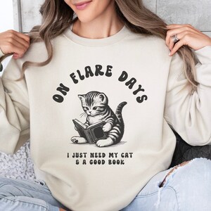 May include: Sand-colored sweatshirt with the text "ON FLARE DAYS" above a black and white illustration of a cat reading a book. Below the illustration is the text "I JUST NEED MY CAT & A GOOD BOOK".