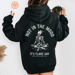 May include: Black hooded sweatshirt with a white graphic of a skeleton sitting in a meditative pose with flames on its head. The text "Not in the mood" is written in a semi-circle above the skeleton and the text "It's flare day" is written below the skeleton. The text "My body chose violence today" is written in smaller font below the text "It's flare day".