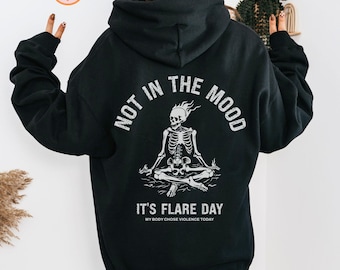 May include: Black hooded sweatshirt with a white graphic of a skeleton sitting in a meditative pose with flames on its head. The text "Not in the mood" is written in a semi-circle above the skeleton and the text "It's flare day" is written below the skeleton. The text "My body chose violence today" is written in smaller font below the text "It's flare day".