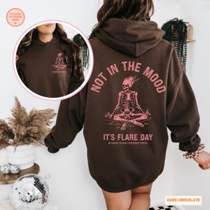 unisex chronic illness hoodie in color dark chocolate. Back design: Graphic of a pink skeleton in flames with a phrase not in the mood its flare day in grunge font in pink. Pocket design: the same skeleton with the text Endometriosis