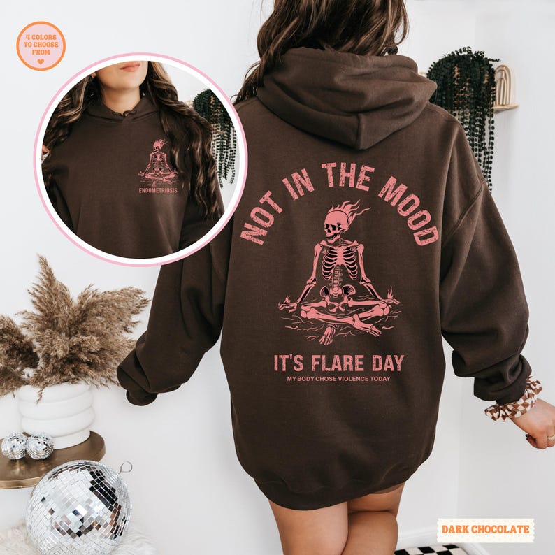 unisex chronic illness hoodie in color dark chocolate. Back design: Graphic of a pink skeleton in flames with a phrase not in the mood its flare day in grunge font in pink. Pocket design: the same skeleton with the text Endometriosis