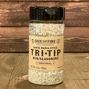 May include: A brown glass jar with a black lid filled with a white and green seasoning blend. The label reads "Oak and Fire Santa Maria Style Tri-Tip Rub/Seasoning *Original* Net Wt: 5.5oz (156g)"