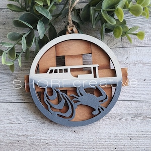 May include: A wooden ornament with a white and gray painted design. The ornament features a boat with a crab and waves in the water. The ornament is perfect for a nautical themed Christmas tree.