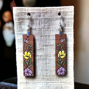 May include: A pair of wooden earrings with a floral design. The earrings are painted with yellow, pink, and green flowers on a brown background.