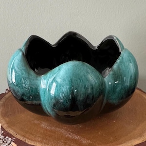 May include: A ceramic bowl with a unique, organic shape. The exterior is a glossy teal green, transitioning to black at the rim and interior. The bowl is placed on a natural wood slice, showcasing its decorative design.