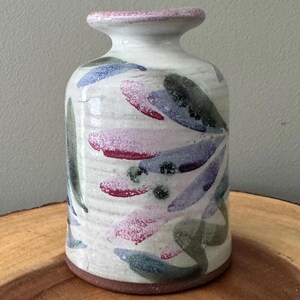 May include: A ceramic vase with a cylindrical body and a narrow neck. The vase is painted with abstract leaf-like shapes in shades of blue, green, pink, and purple on a white background. The rim is a light pink color, and the base is brown.