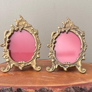May include: Two ornate, antique gold-colored picture frames with oval openings and red backgrounds. The frames have intricate detailing and stand on small feet. Each frame has a decorative element at the top.