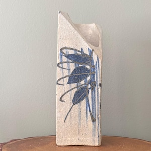 May include: A rectangular ceramic vase with a cream-colored base and blue leaf and stripe design. The vase has a unique angled top and is displayed on a wooden surface.