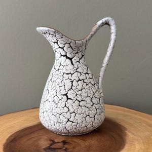 May include: A small, white ceramic pitcher with a crackle glaze pattern and a curved handle. The pitcher has a narrow neck and a slightly flared spout. The crackle pattern is black, creating a striking contrast against the white background. The pitcher is likely vintage.