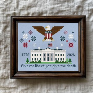 May include: Framed cross-stitch art with a light blue background, depicting the White House, an eagle, and snowflakes. The design includes the years 1776 and 2026, and the text "Give me liberty or give me death."