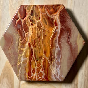May include: Hexagonal ceramic coaster with an abstract design. The coaster features a blend of warm colours, including shades of orange, gold, and red, creating a marbled effect. The design has a glossy finish and is suitable for home decor.