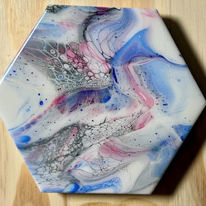 Puede incluir: Azulejo de cerámica hexagonal con un diseño abstracto de arte fluido. El azulejo presenta una mezcla de tonos blancos, rosas, azules y grises, con patrones intrincados y formaciones celulares. La obra de arte está sobre un fondo de vetas de madera clara.