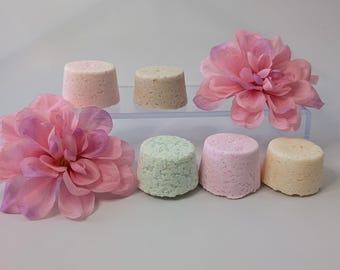 Puede incluir: Un surtido de seis bombas de baño cilíndricas de colores en tonos rosa, verde y beige, dispuestas con dos grandes flores de tela rosa. Las bombas de baño están sobre una estantería transparente.