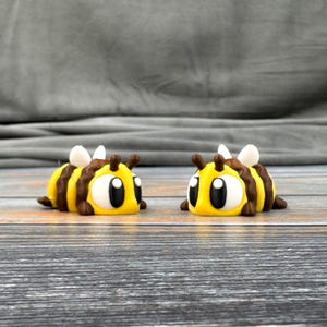 May include: Two small, yellow and brown bee figurines with black eyes and white highlights. The bees are facing each other on a wooden surface.