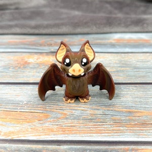 May include: A small brown bat figurine with large black eyes, a light tan nose, and pointed ears with tan inner details. The bat has outstretched wings and small feet, featuring a smooth, molded texture throughout its body and facial features.