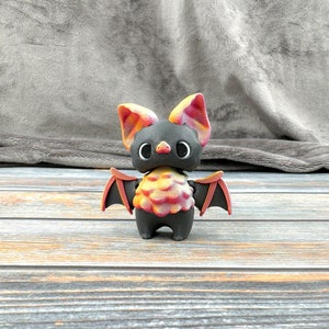 May include: A small, black bat figurine with red and yellow wings. The bat has large, round eyes and a cute expression.