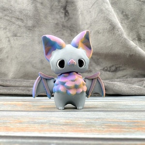May include: A small, grey, 3D printed bat figurine with iridescent purple and blue accents. The bat has large, black eyes and a textured, bumpy body.