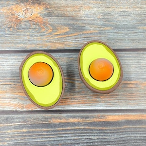 May include: Two wooden avocado halves with yellow interiors and orange pit centers. The avocado halves are on a wood background.