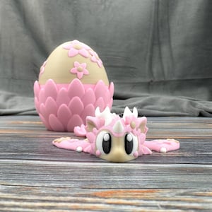 May include: A pink and beige decorative egg with flower accents and a matching pink dragon figurine. The egg has a textured, petal-like base. The dragon has large black eyes and white accents.