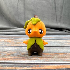 May include: A 3D-printed figurine of a pumpkin-headed character. The figure has an orange pumpkin head with large black eyes, a green leafy stem, and a brown body. The character has small orange arms and brown feet. The figurine is set against a gray background.