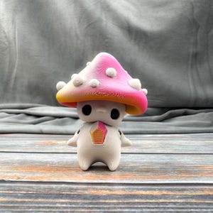 May include: A whimsical mushroom figurine with a pink and yellow cap, adorned with white spots. The figurine has a white body, black eyes, and small arms. A colorful design is on the chest. The mushroom is a unique decorative item.