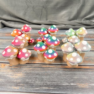 May include: A collection of colorful mushroom figurines with white polka dots on the caps. The mushrooms have a variety of colors, including pink, orange, green, and blue. They are arranged on a wooden surface.