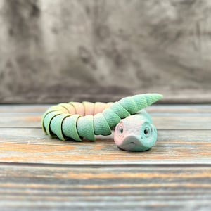 Peut inclure: Une petite figurine de serpent articulée présentant un dégradé de couleurs passant du rose pastel doux au vert menthe. Le jouet possède un corps segmenté et flexible ainsi qu'une tête détaillée avec des yeux visibles, en position enroulée.