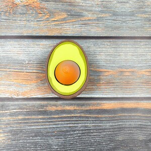 May include: A wooden avocado-shaped toy with a yellow avocado pit inside. The toy is on a wooden surface.