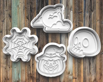 May include: Four white plastic Halloween cookie cutters: a pumpkin with a witch's hat, a skull, a spider web, and a crescent moon with a bat.