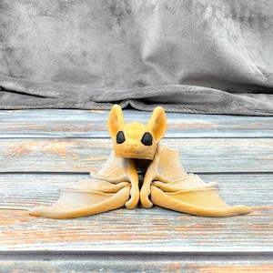 May include: A tan-coloured bat figurine with large, pointed ears and solid black eyes. The bat is depicted in a resting position with its wings folded neatly against its body, showcasing a detailed, layered texture across the surface of the wings.