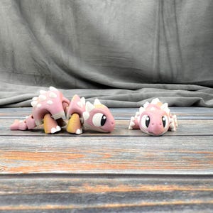 May include: Two pink and white 3D printed dinosaur toys with large eyes. The dinosaur on the left is walking on all fours, while the dinosaur on the right is lying on its belly.