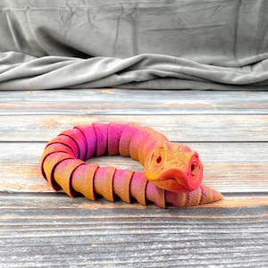 Peut inclure: Un jouet serpent articulé imprimé en 3D avec un dégradé de couleurs vives passant du magenta et violet profond à l'orange et jaune éclatants. Le corps segmenté est composé de multiples pièces imbriquées les unes dans les autres.