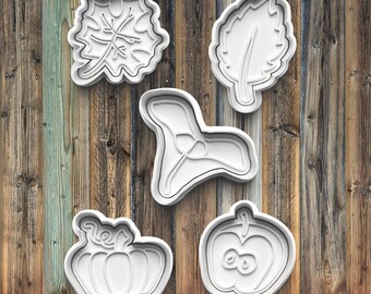 May include: Five white plastic cookie cutters in the shapes of a leaf, a large leaf, a flower, a pumpkin, and an apple with an eight-shaped core. The cutters are arranged on a wooden background.
