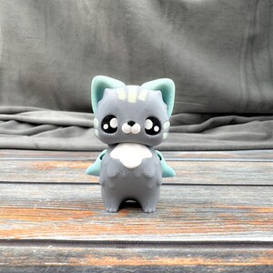 May include: A gray cat figurine with large, expressive eyes and light blue accents on the ears and wings. The cat has a white chest and a small, black nose. The figurine is standing on a wooden surface.