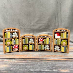 May include: Four miniature wooden bookshelves with various items. Each shelf is filled with colorful books, a mushroom, a frog, and a Santa hat. The shelves are brown with decorative scrollwork at the top.