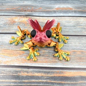 May include: A 3D printed articulated frog figure featuring a gradient color scheme of orange, yellow, and green on its body and limbs. The frog has two large, solid black eyes and two prominent, upright, dark pink leaf-like structures on its head.
