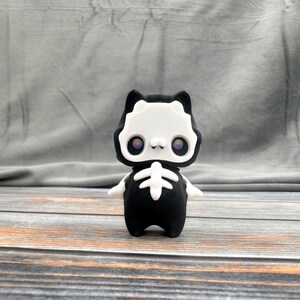May include: A 3D-printed figurine of a cat in a skeleton costume. The cat is black and white with a rib cage design on the front. The eyes are a vibrant purple. The figurine is standing on a wooden surface.